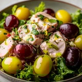 A bowl of food with chicken, grapes, and olives.